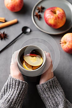 Hands hold a gray glass with homemade apple punch or cider with apples and cinnamon on a dark background with fresh fruits and