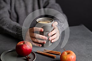 Hands hold a gray glass with homemade apple punch or cider with apples and cinnamon on a dark background with fresh fruits and