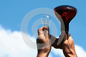 Hands hold infrared lamp and transparent incandescent light bulb against blue sky background