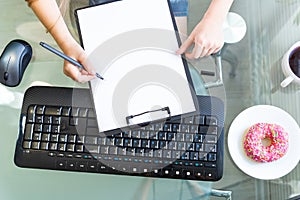 Hands hold a pen over a blank white sheet of paper, keyboard, computer mouse, donut on a plate and coffee. Home office