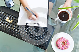 Hands hold a pen over a blank white sheet of paper, keyboard, computer mouse, donut on a plate and coffee. Home office