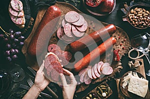 Hands hold sandwich with slices of salami above a wooden cutting board on dark background. Meat appetizer with sausage, ham,