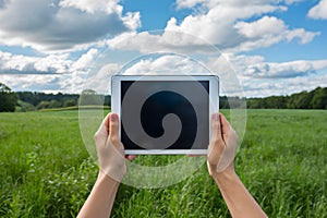 Hands hold tablet against a lush green field with distant trees photo