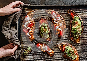 Hands holding baking tray with baked sweet potato toasts with chickpeas, tomatoes, goat cheese, sauce guacamole, avocado,