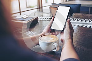 Hands holding black mobile phone with blank desktop screen with coffee cup on wooden table in cafe