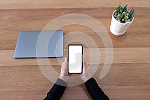 Hands holding black mobile phone with blank desktop screen with laptop and cactus pot on wooden table background