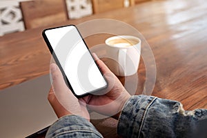 Hands holding black mobile phone with blank desktop screen with laptop and coffee cup on the table