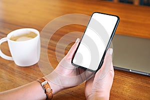 Hands holding black mobile phone with blank desktop screen with laptop and coffee cup on the table