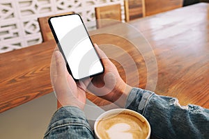 Hands holding black mobile phone with blank desktop screen with laptop and coffee cup on the table