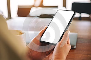 Hands holding black mobile phone with blank desktop screen with laptop and coffee cup on the table