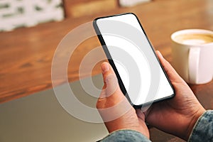 Hands holding black mobile phone with blank desktop screen with laptop and coffee cup on the table