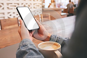 Hands holding black mobile phone with blank desktop screen with laptop and coffee cup on the table