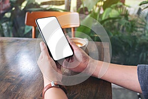 Hands holding black mobile phone with blank desktop screen with laptop and coffee cup on the table