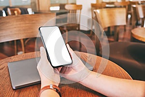 Hands holding black mobile phone with blank desktop screen with laptop computer on the table