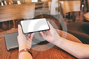 Hands holding black mobile phone with blank desktop screen with laptop computer on the table