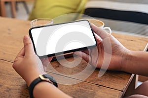 Hands holding black mobile phone with blank screen horizontally with coffee cup on wooden table in cafe