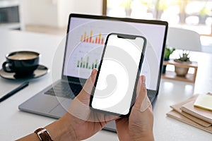 Hands holding a black mobile phone with blank screen while using laptop computer in office