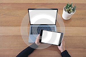 Hands holding a black tablet and laptop with blank white screen on wooden table in office