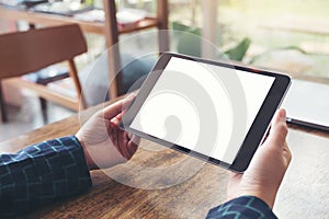 Hands holding black tablet pc with blank white desktop screen and laptop on wooden table in cafe