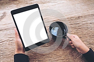 Hands holding black tablet pc with blank white screen and coffee cup on vintage wooden table in cafe