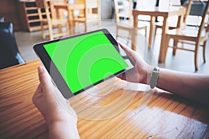 Hands holding black tablet pc with white blank green screen on wooden table in modern cafe