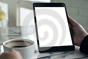 Hands holding black tablet pc with white blank screen and coffee cup on table background