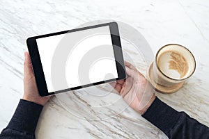 Hands holding black tablet pc with white blank screen and coffee cup on table background