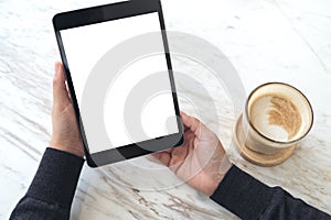 Hands holding black tablet pc with white blank screen and coffee cup on table background