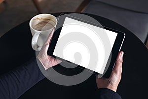 Hands holding black tablet pc with white blank screen and coffee cup on table background