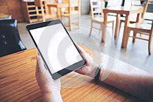 Hands holding black tablet pc with white blank white screen on wooden table in modern cafe