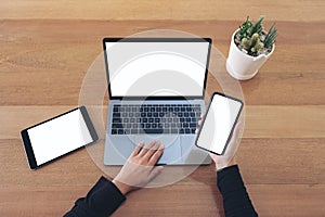 Hands holding a blank white screen mobile phone and laptop with black tablet on wooden table in office
