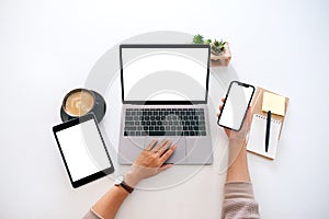 Hands holding a blank white screen mobile phone with laptop computer and tablet pc on the table in office