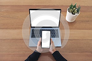 Hands holding a blank white screen mobile phone and laptop on wooden table in office