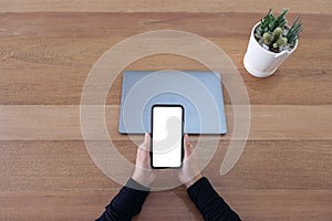 A hands holding a blank white screen mobile phone and laptop on wooden table in office