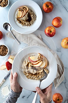 Hands holding a bowl of steel cut oats with brown sugar and sliced apple above a breakfast table top in soft focus.