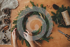 Hands holding christmas wreath with fir branches, berries, pine cones, and thread, scissors on rural wooden table. Rustic