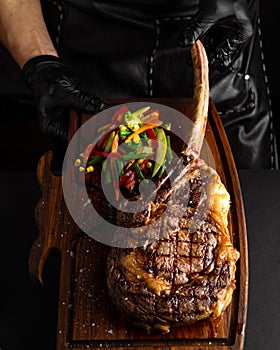 Hands holding cooked Tomahawk steak on a serving board. Low key image, vertical orientation