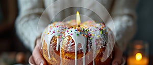 Hands holding Easter cake Kulich decorated with white dripping icing, colored sprinkles and burning candle. Blurred