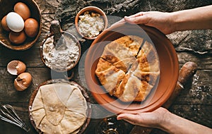 Hands holding flat bread or round pies with cottage cheese, eggs, herb in plate on rustic wooden background. Homemade baking,