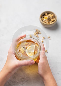 Hands holding a glass cup with chamomile herbal tea on a light background with dry flowers. The concept of a healthy drink for