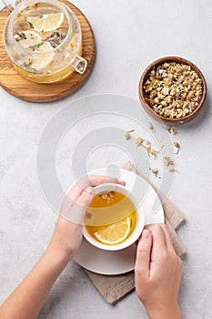 Hands holding a glass cup with chamomile herbal tea on a light background with dry flowers and teapot. The concept of a healthy