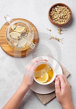 Hands holding a glass cup with chamomile herbal tea on a light background with dry flowers and teapot. The concept of a healthy