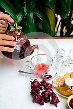 Hands holding glass teapot with hibiscus tea with cups and crystalized fruits and jam on a table