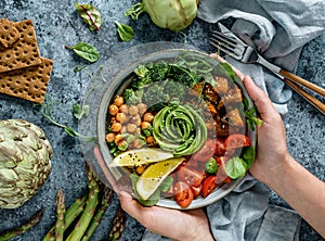 Hands holding healthy superbowl or Buddha bowl with salad, baked sweet potatoes, chickpeas, broccoli, hummus, avocado, sprouts on