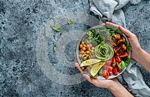 Hands holding healthy superbowl or Buddha bowl with salad, baked sweet potatoes, chickpeas, broccoli, hummus, avocado, sprouts on