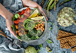 Hands holding healthy superbowl or Buddha bowl with salad, baked sweet potatoes, chickpeas, broccoli, hummus, avocado, sprouts on