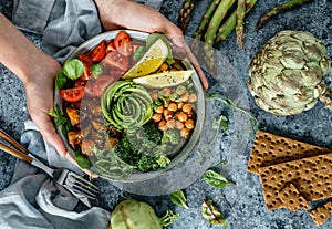 Hands holding healthy superbowl or Buddha bowl with salad, baked sweet potatoes, chickpeas, broccoli, hummus, avocado, sprouts on photo