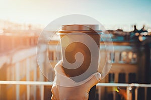 Hands holding hot craft cup of coffee or tea in morning sunlight with view to blurred city background. Enjoy, lifestyle