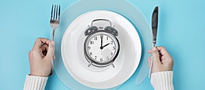 Hands holding knife and fork above alarm clock on white plate on blue background. Intermittent fasting, Ketogenic dieting, weight