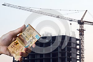 Hands holding money tenge on the background of the house under construction. Buying a new apartment in Kazakhstan for tenge. New